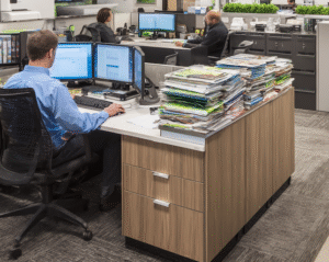 Oficina moderna con empleados trabajando entre ordenadores y grandes pilas de documentos. La imagen representa el entorno corporativo donde se generan residuos electrónicos y la importancia de toda auditoría de reciclaje de RAEE en Navarra para cumplir con la normativa RAEE.