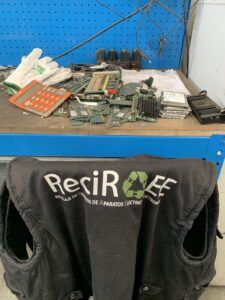 Mesa de trabajo con componentes electrónicos en desuso como placas, discos duros, memorias y cables, junto a herramientas y un chaleco de ReciRAEE en primer plano, imagen que muestra la gestión real de residuos electrónicos (RAEE), su clasificación y tratamiento profesional en un entorno de trabajo.
