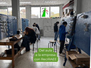 Aula-taller en Navarra con personas trabajando en paneles y mesas de prácticas, en una actividad vinculada a la gestión de residuos electrónicos, con el texto “Del aula a la empresa con ReciRAEE”, imagen que muestra la conexión entre formación práctica, clasificación de RAEE y aprendizaje del trabajo en una empresa autorizada.